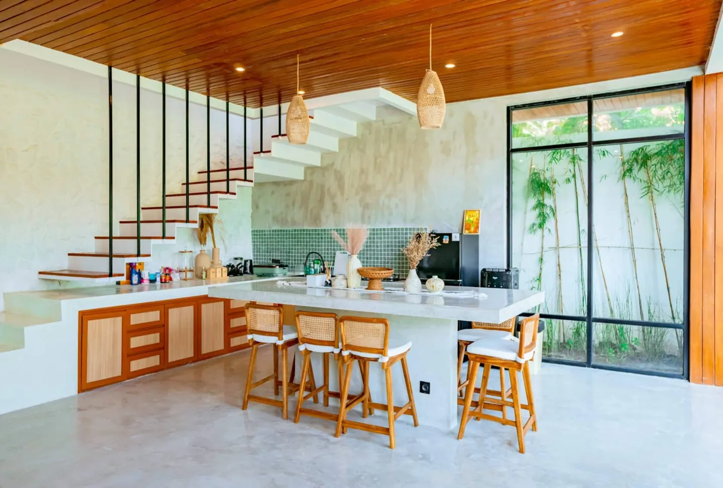 Fully equipped kitchen with floating concrete stairs, a large concrete island, rattan bar stools and a green mosaic backsplash